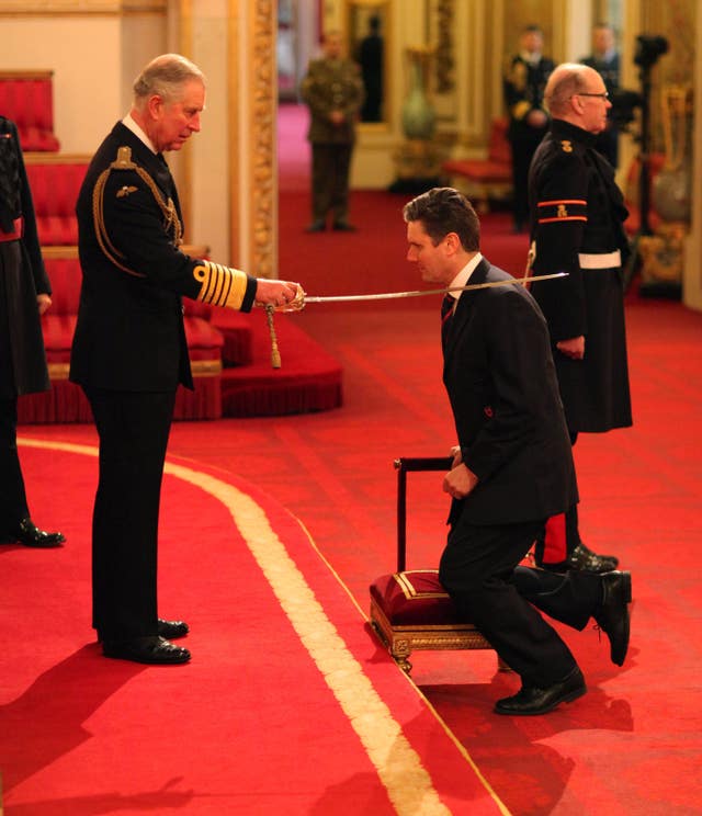 Prince Charles, who is in military attire and is holding a sword, knights Keir Starmer, who is kneeling on a cushion. They are at a hall in Buckingham Palace.