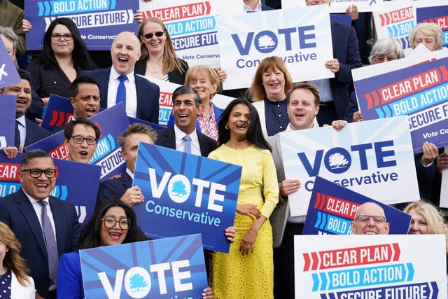 Rishi Sunak in a suit and his wife Akshata Murty in a yellow dress surrounded by campaigners holding 'vote Conservative' signs