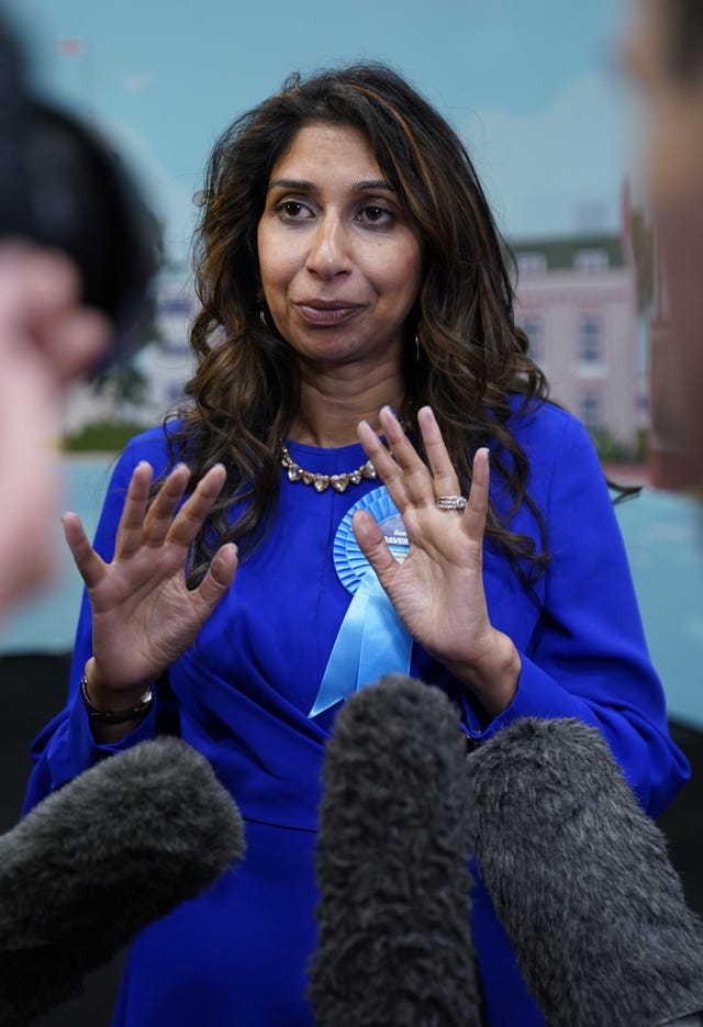 Suella Braverman in blue top with pale blue party rosette with her hands raised