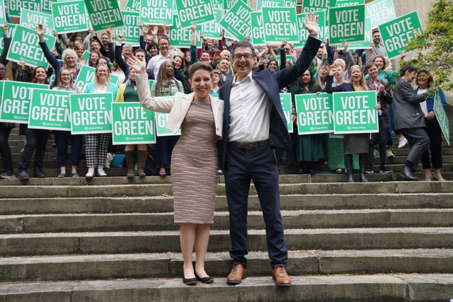 Carla Denyer and Adrian Ramsay wave as they stand together on a set of steps in front of supporters holding placards saying 'Vote Green'