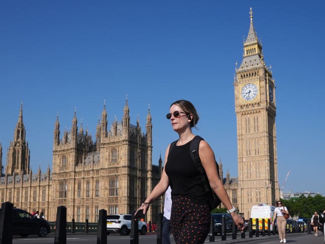 A woman wearing sunglasses walks past Big Ben as the sun shines in a blue, cloudless sky
