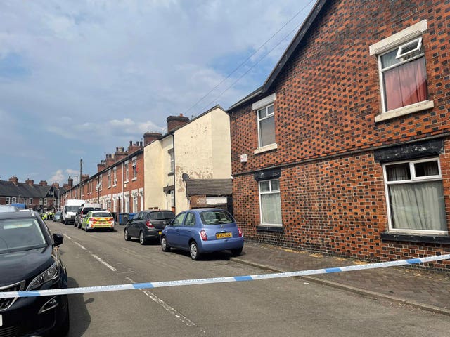 A street of terraced houses. There are cars parked on the road but it is taped off with police tape. Police cars and police officers are visible in the distance at the bottom of the street