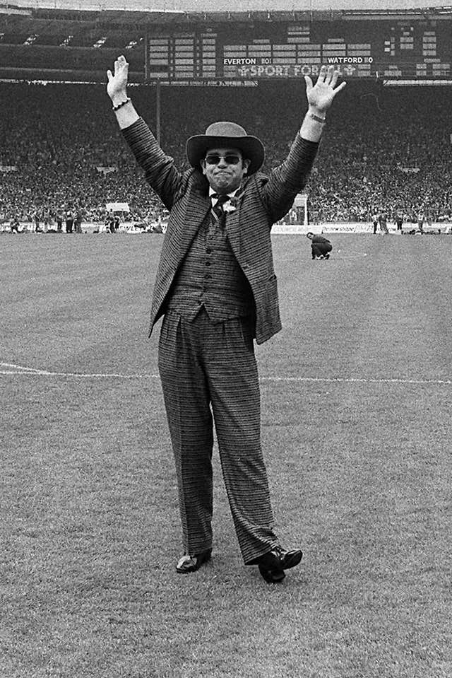 A black and white photo of Elton John waving to a crowded stadium