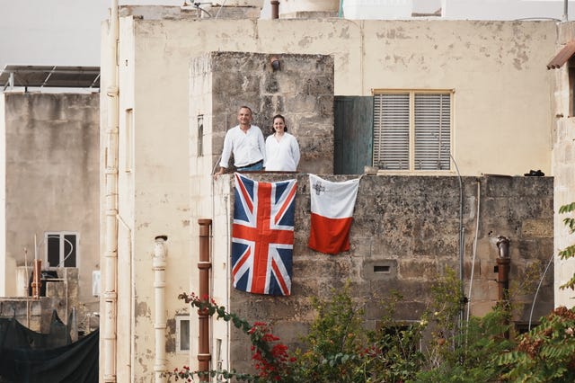 Two people standing on a balcony with Union and Maltese flags draped over it