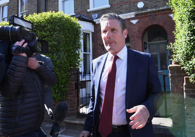 Keir Starmer outside the gate of his home in north London. Sir Keir is wearing a navy suit with a red tie, and is next to a cameraman filming him.