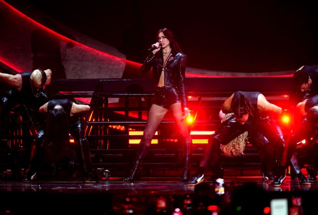 Dua Lipa dressed in leather jacket and shorts while singing on a stage with dancers