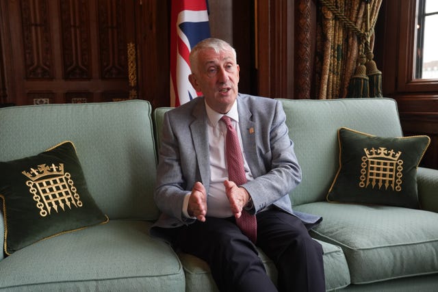 Sir Lindsay Hoyle gestures while sitting on a couch between two parliamentary cushions