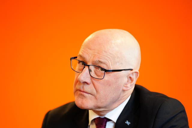 Headshot of John Swinney, seated in front of an orange wall