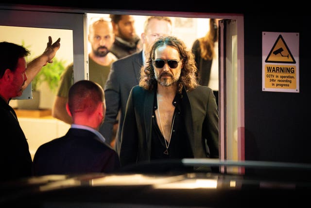 Russell Brand wearing dark glasses walking through a doorway to a waiting car.
