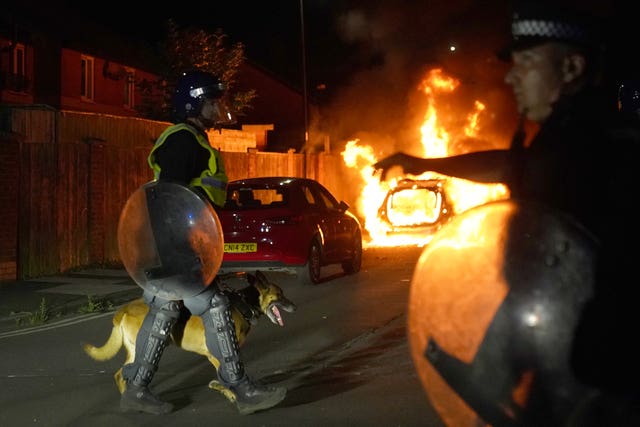 Police officers in riot gear, one with an alsatian on a leash, walk past a burning police car during riots in Hartlepool