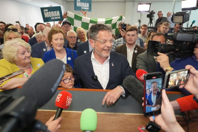 A man with a grey suit and clear, thick rimmed glasses, smiles for people taking photos on smartphones. There are a number of microphones pointed his way and he is surrounded by supporters and cameras.