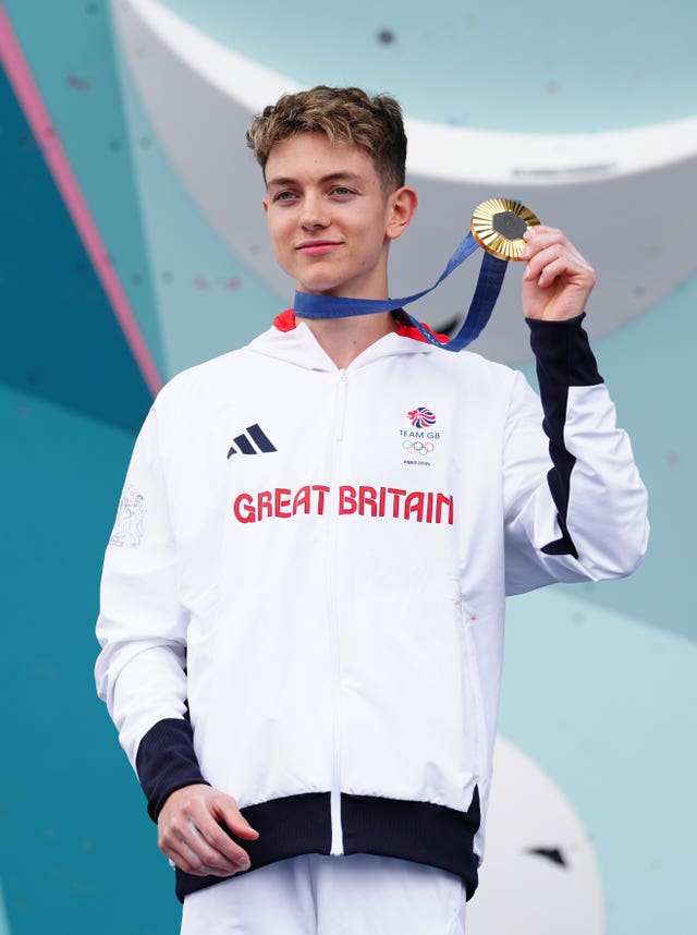 Toby Roberts in white Team GB tracksuit holding up his gold medal