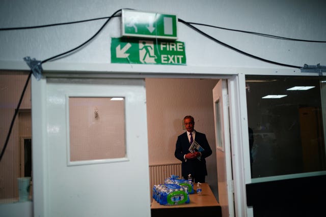 Nigel Farage pictured through an open door in a hall behind a folding table with bottles of water on it. A fire exit sign and cables are prominent over the open door