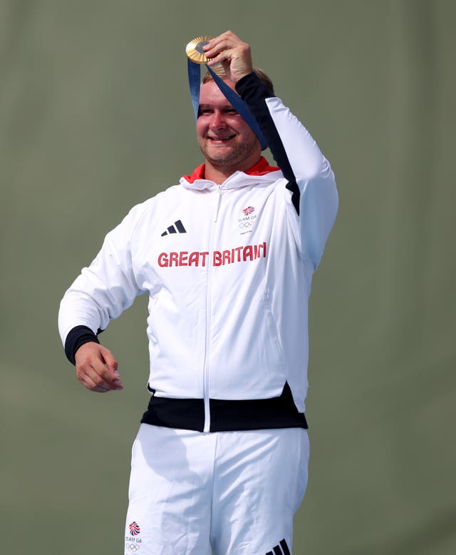 Nathan Hales, dressed in a white Team GB tracksuit, holds his gold medal above his head.