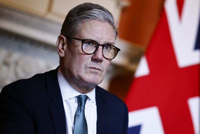 Sir Keir Starmer with a stern look on his face standing nearby a Union flag