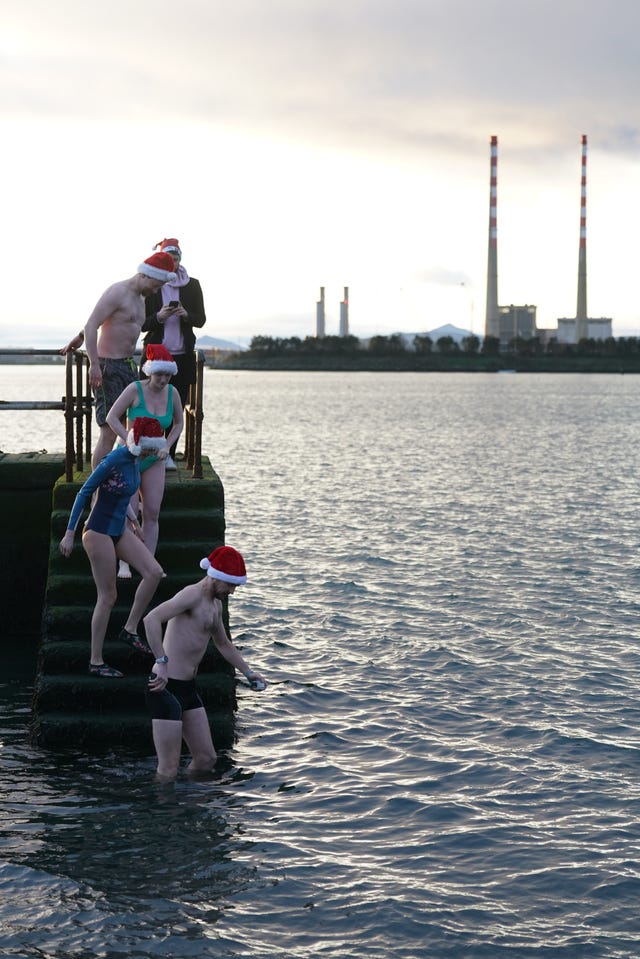 Four swimmers in Santa hats climbing down concrete steps into the sea with industrial chimneys behind them 