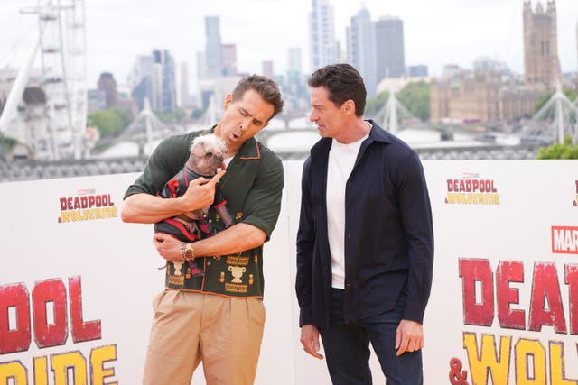 Ryan Reynolds in a green shirt and beige trousers and Hugh Jackmand in a blue shirt and jeans hold Peggy, a pug dressed in a deadpool costume. Behind them are posters for Deadpool & Wolverine and the London cityscape.