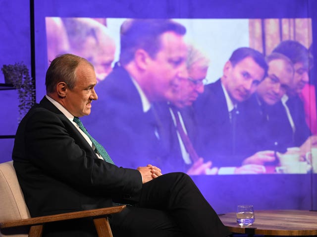 Liberal Democrats leader Ed Davey sitting in front of a screen showing an image from when he was in the coalition cabinet with David Cameron and Jeremy Hunt