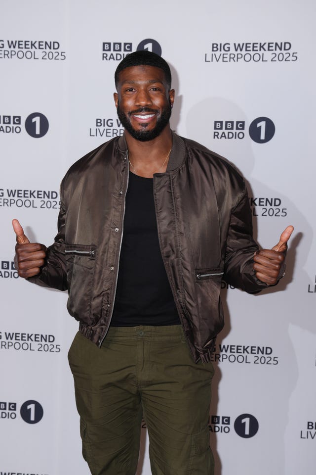 Toby Olubi smiling and giving a thumbs up with both hands, standing in front of Radio 1 signage