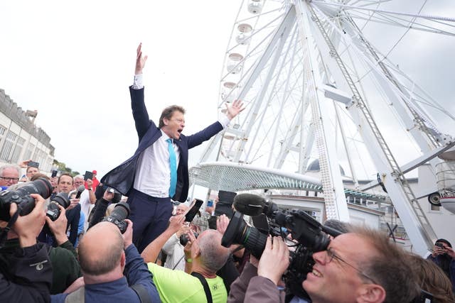 Richard Tice stands above photographers with his arms in the air beside a white big wheel ride