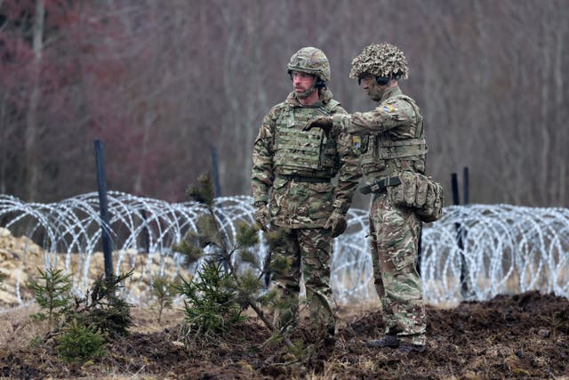 Prince of Wales in Army fatigues, standing next to a soldier in a field, with barbed wire behind him