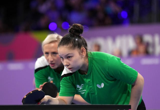 Anna Hursey crouches at the table tennis table, dressed in green, waiting for a serve. 