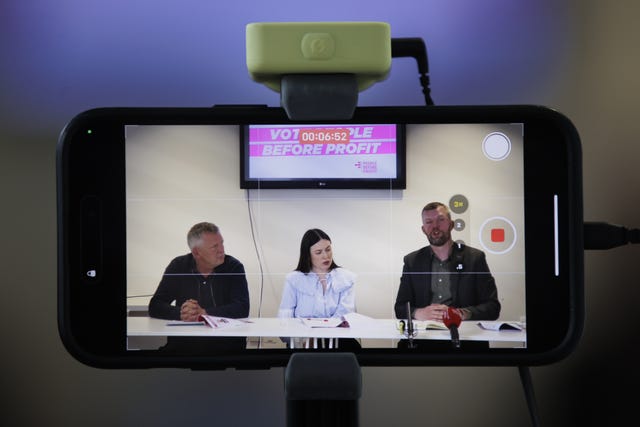 An iphone screen shows video still of three people sat at a table, a TV screen behind them