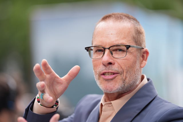 Guy Pearce smiling with one hand in the air wearing glasses, a brown shirt and blue suit jacket