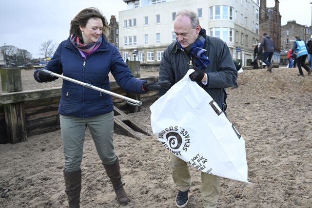 Sir Ed Davey holding a bag while a woman puts litter in it, on a beach