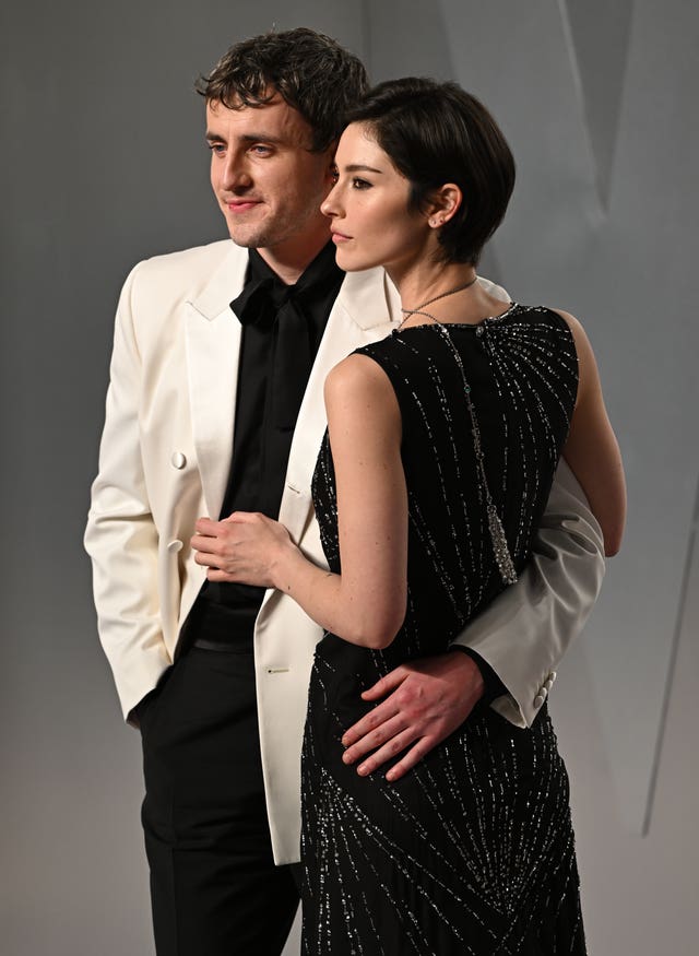 Paul Mescal, wearing a white blazer over a black shirt and black trousers, standing with his arm around Gracie Abrams' waist. Abrams is wearing a black clingy dress with sparkling beaded silver stripes and has her hand resting on Mescal's torso.