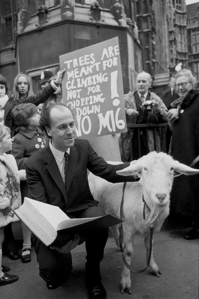 Norman Tebbit on his knees with a petition in his right hand while stroking a goat with his left hand in front of placard-holding campaigners