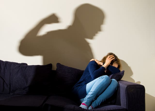 Posed picture of a woman cowering on a sofa with the shadow of a man brandishing his fist cast over her.