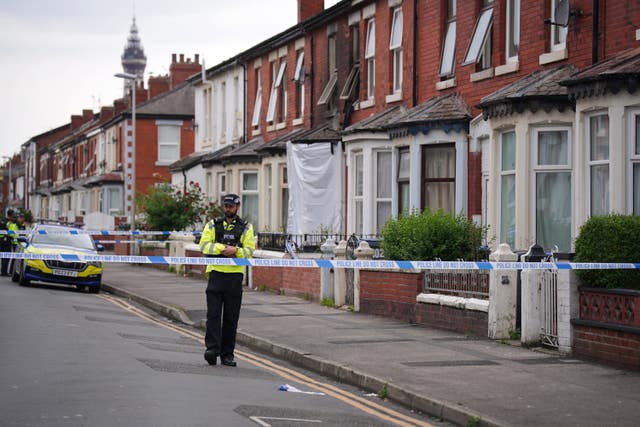 Police tape cordons off a street, a police man stands in the road and one house is charred and burned