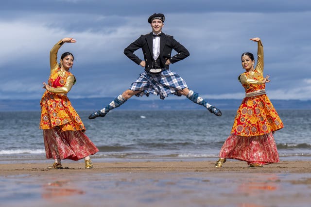 A Scottish dancer in a blue kilt jumps in the air with his legs apart in between two Bollywood dancers doing symmetrical poses