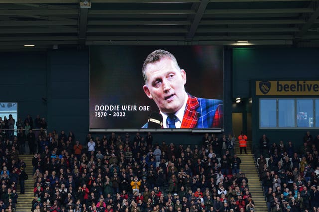 A screen featuring a picture of Doddie Weir with the caption 'Doddie Weir OBE 1970-2022' on display within a rugby stadium filled with fans