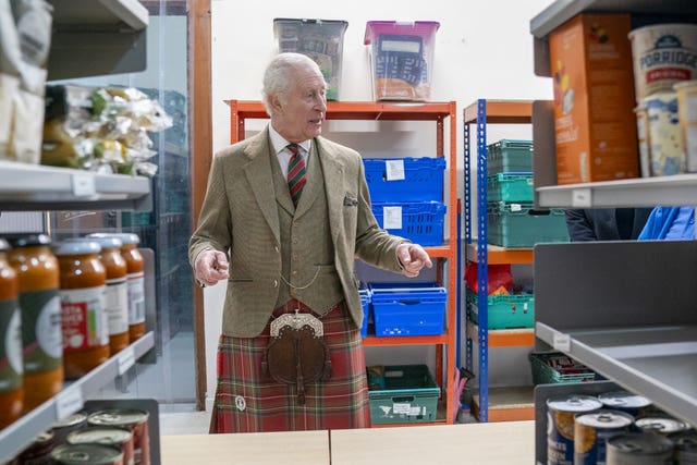 The King dressed in a kilt in an aisle, with food on the shelves beside him