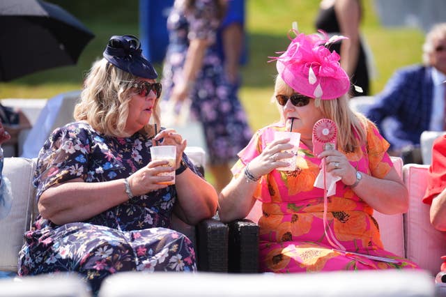 Two women wearing outlandish hats and glamourous dresses sip from drinks at Royal Ascot