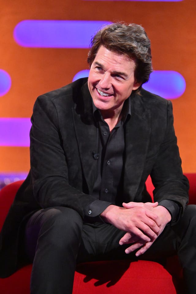 Tom Cruise smiling with his hands crossed, sitting on a red couch and wearing a black suit