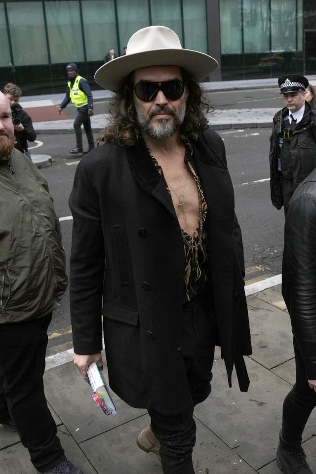 Russell Brand walking outside court, wearing a coat and shirt, which is unbuttoned to the middle, a white hat and sunglasses