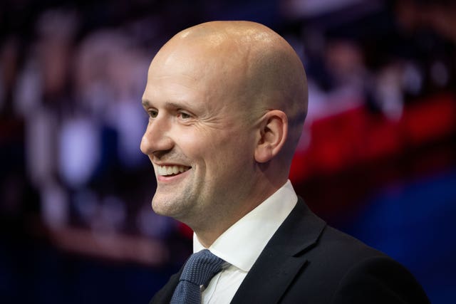 Head shot of SNP Westminster leader Stephen Flynn, wearing a dark suit jacket, white shirt and dark blue tie, looking to the left and smiling