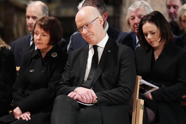 John Swinney in black suit, seated alongside his wife and in front of Kate Forbes