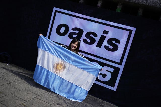 An Oasis fan holding an Argentina flag poses for a photo in front of an Oasis Live 25 sign 