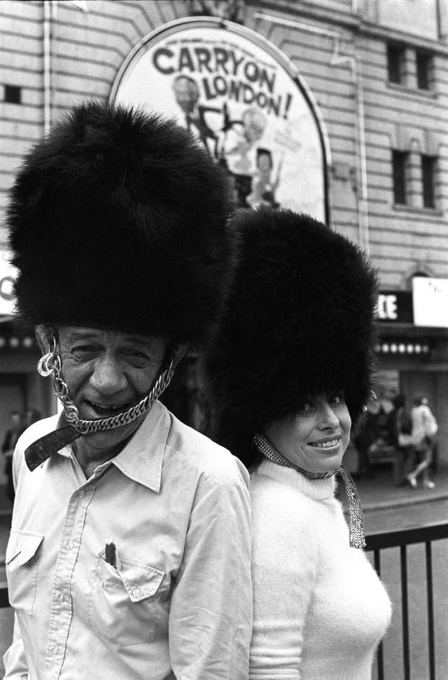Sid James and Barbara Windsor pictured outside the Victoria Palace Theatre wearing busbies (PA)