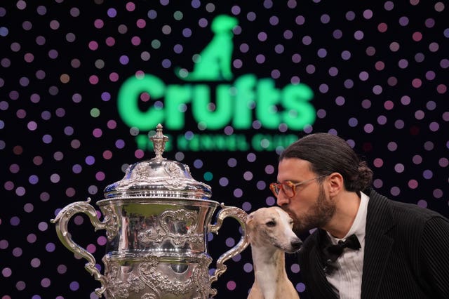 A man kissing a whippet in front of a Crufts sign besides the winning trophy