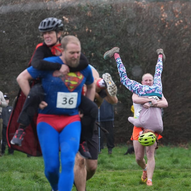 A man in the foreground wearing a Superman costume carrying his wife on his back while a man in the background carries his wife upside down in front of him