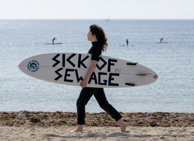 A protesting surfer walks on the beach with a surf board reading 'sick of sewage'