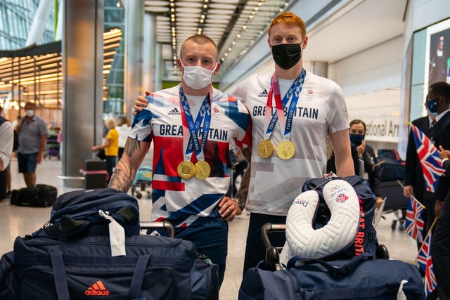 Adam Peaty and Tom Dean, wearing masks and carrying luggage, pose for a picture wearing their Olympic medals