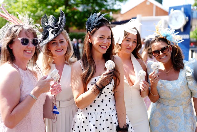 Five women dressed elegantly during day three of Royal Ascot holding electric hand fans to their faces