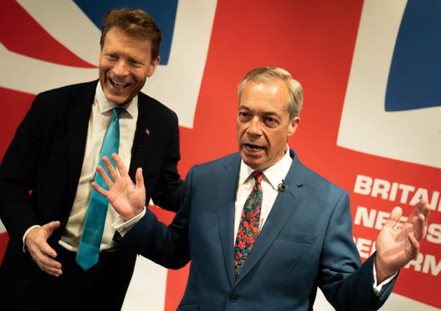 Richard Tice laughing while Nigel Farage speaks, with his hands raised, in front of a British flag banner