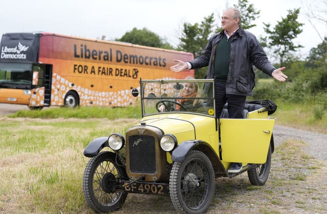 Liberal Democrats leader Sir Ed Davey stands up in the passenger seat of a small, yellow open-top classic car, with Liberal Democrat candidate for Chippenham Sarah Gibson sat down in the driving seat, with the Lib Dem battlebus in the background, during a visit to Owl Lodge in Lacock, Wiltshire, while on the General Election campaign trail